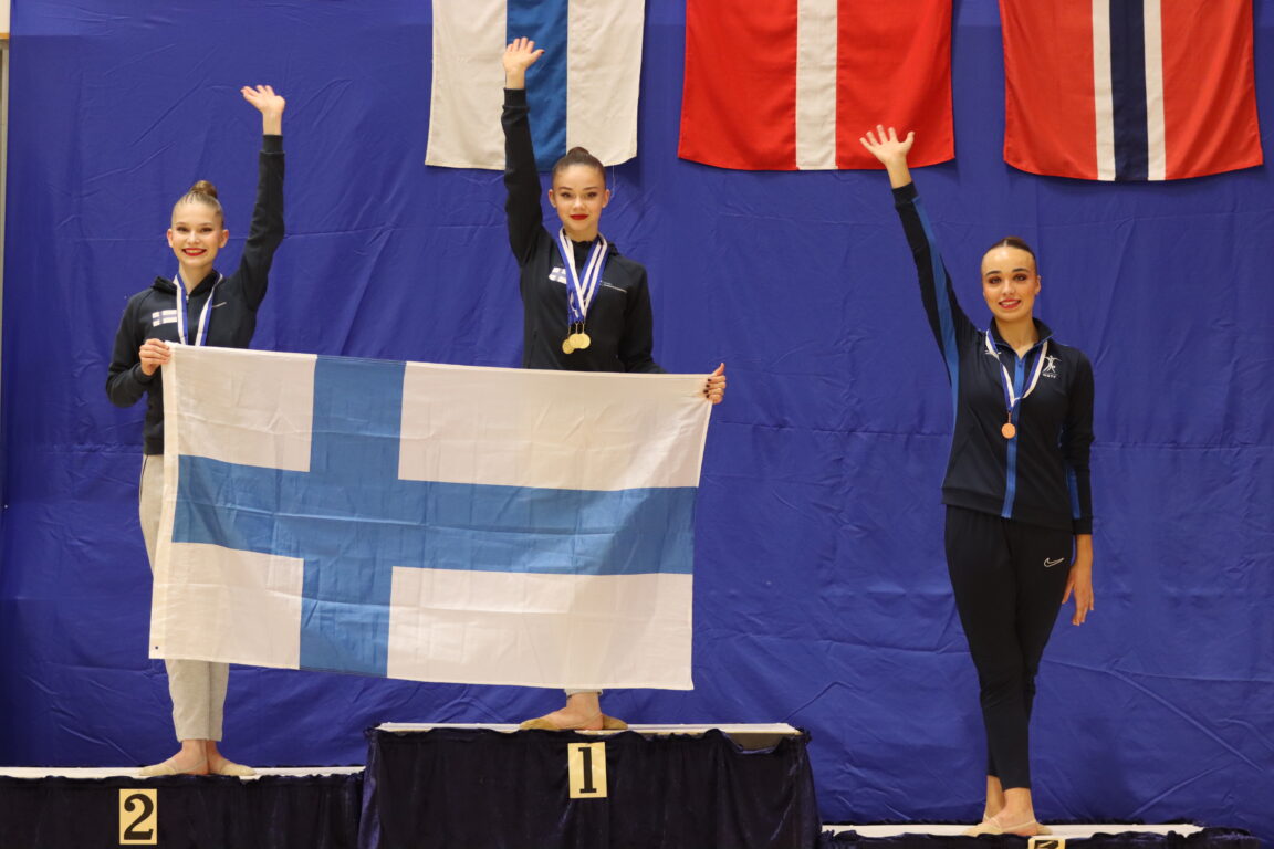 Rytmisiä voimistelijoita palkintopallilla. Voimistelijat vilkuttavat. Korkeimmalla pallilla keskellä on Emmi Piiroinen ja vasemmalla hopeapallilla on Lia Kallio. He pitävät edessään Suomen lippua.