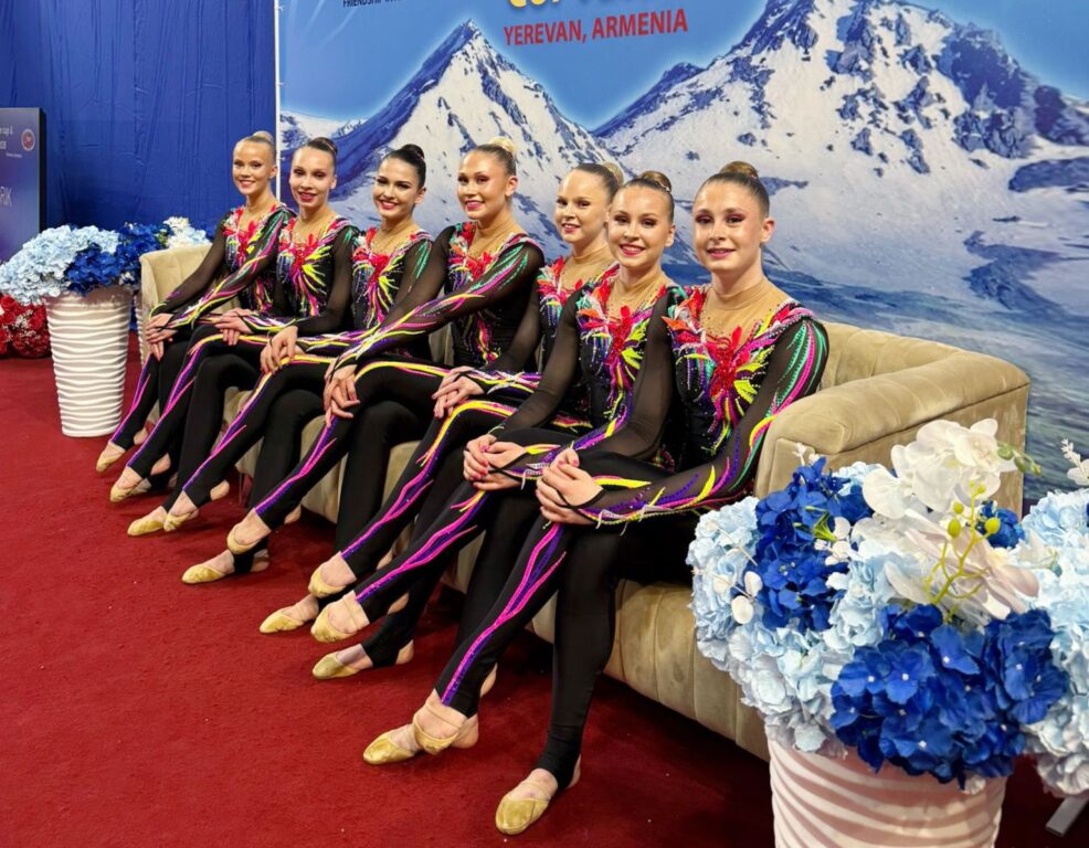 Gloria Gloria-joukkue istuu vaalealla sohvalla kisa-asuissaan, taustalla vuoren kuva.